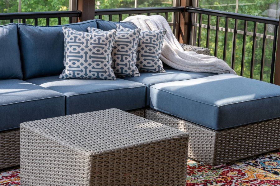 The Ensley 55+ Luxury Apartments with Blue outdoor sectional with patterned pillows, a knit blanket, and a wicker table on a colorful rug.