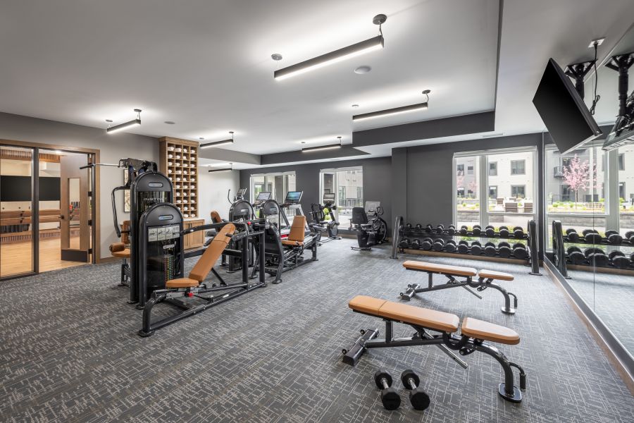 The Ensley 55+ Luxury Apartments with Modern gym with weight machines, benches, dumbbells, cardio equipment, large mirrors, and windows showing buildings outside.