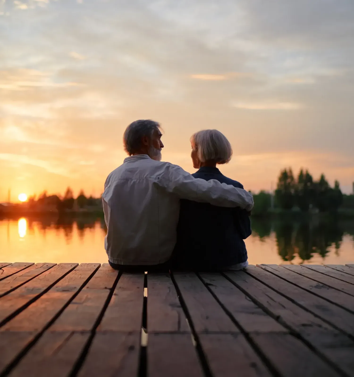 The Ensley 55+ Luxury Apartments with An older couple sits on a wooden dock, watching the sunset over a calm lake.