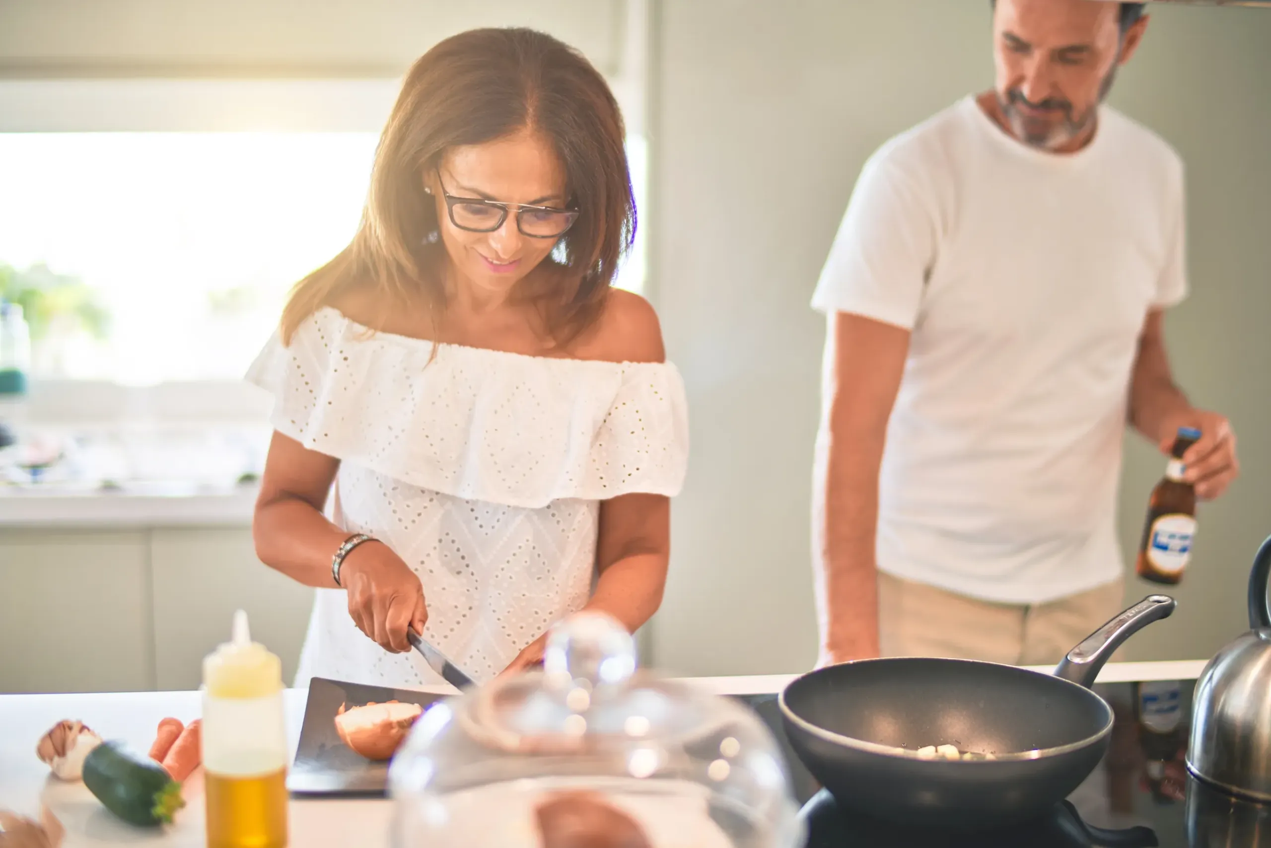 The Ensley 55+ Luxury Apartments with Woman chopping vegetables while a man stands nearby holding a beer in a bright kitchen.