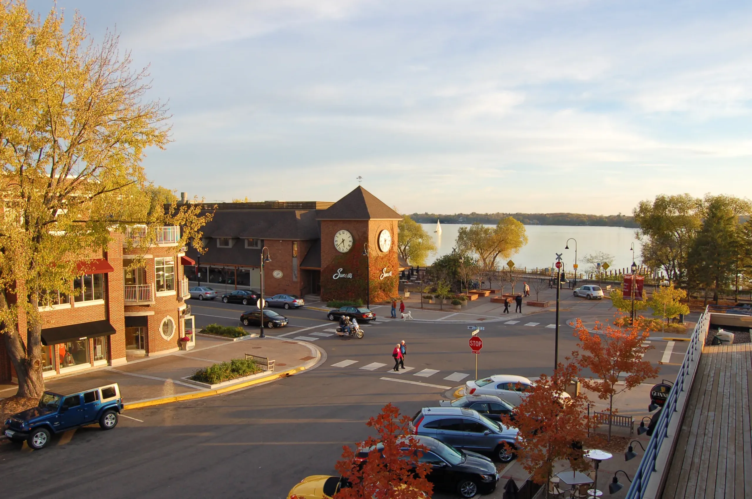 The Ensley 55+ Luxury Apartments with Outdoor view of a lakeside town square with a clock tower, parked cars, and people crossing the street.