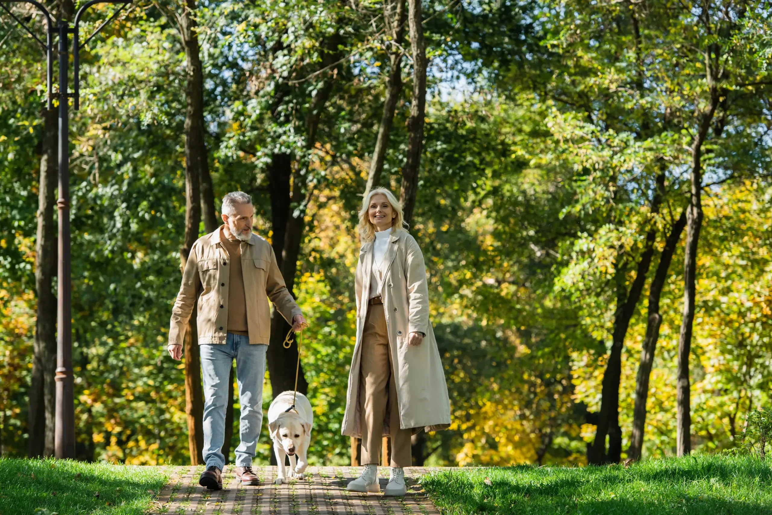 The Ensley 55+ Luxury Apartments with Two people walking a dog together on a sunny path in a green, tree-filled park.