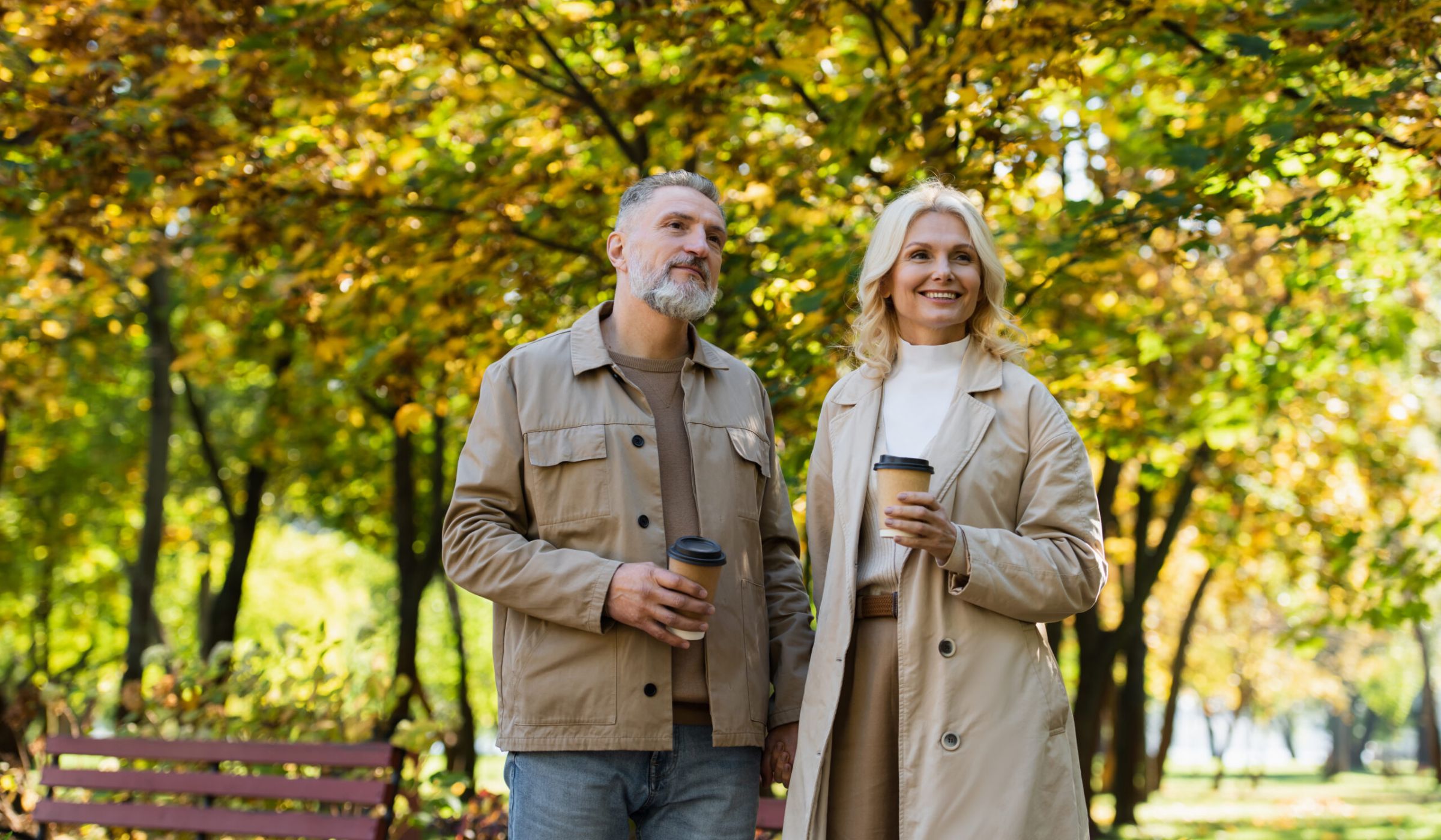 The Ensley 55+ Luxury Apartments with Smiling couple stands outdoors in a park with coffee cups, surrounded by autumn trees and sunlight.