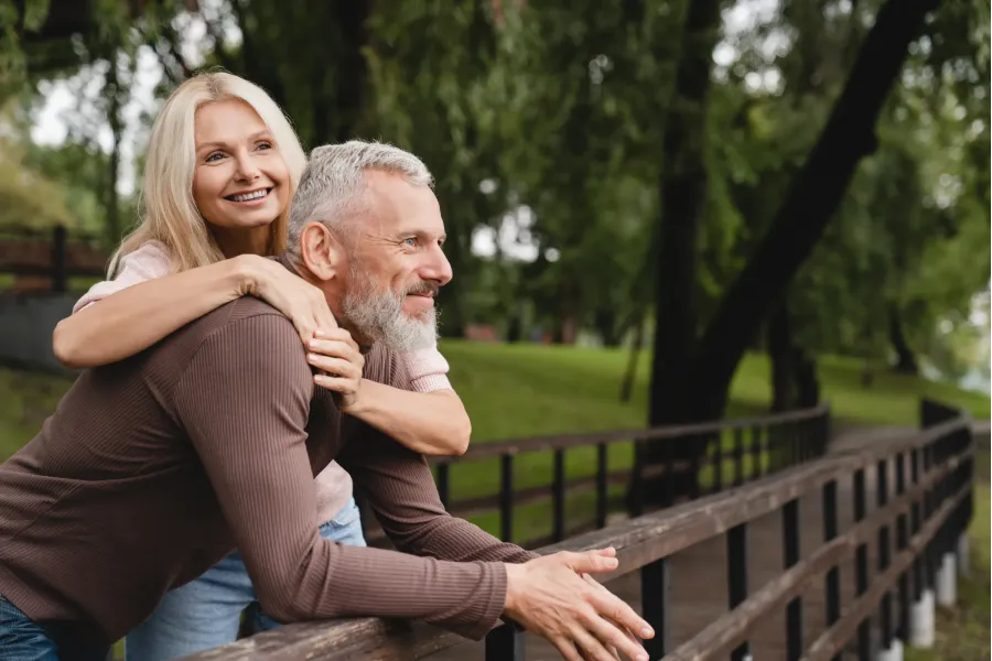The Ensley 55+ Luxury Apartments with Smiling older couple embraces on a wooden bridge in a green, tree-filled park.