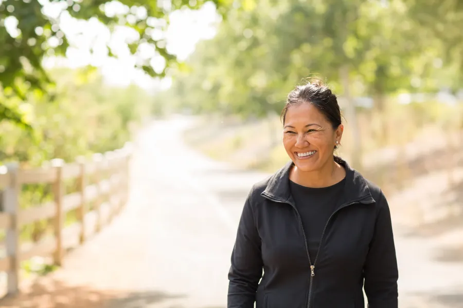 The Ensley 55+ Luxury Apartments with Woman smiling outdoors on a sunny day, standing on a path surrounded by trees and greenery.