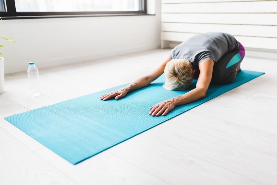 The Ensley 55+ Luxury Apartments with Person doing a child’s pose yoga stretch on a blue mat indoors, with a water bottle nearby.