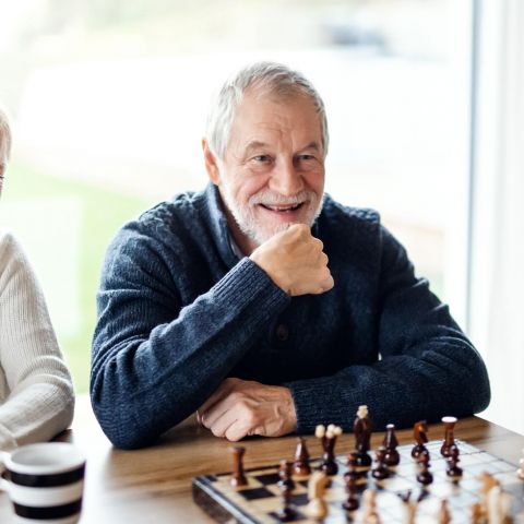 The Ensley 55+ Luxury Apartments with Two smiling older adults watch a chess game, seated at a table with coffee cups and a chessboard.