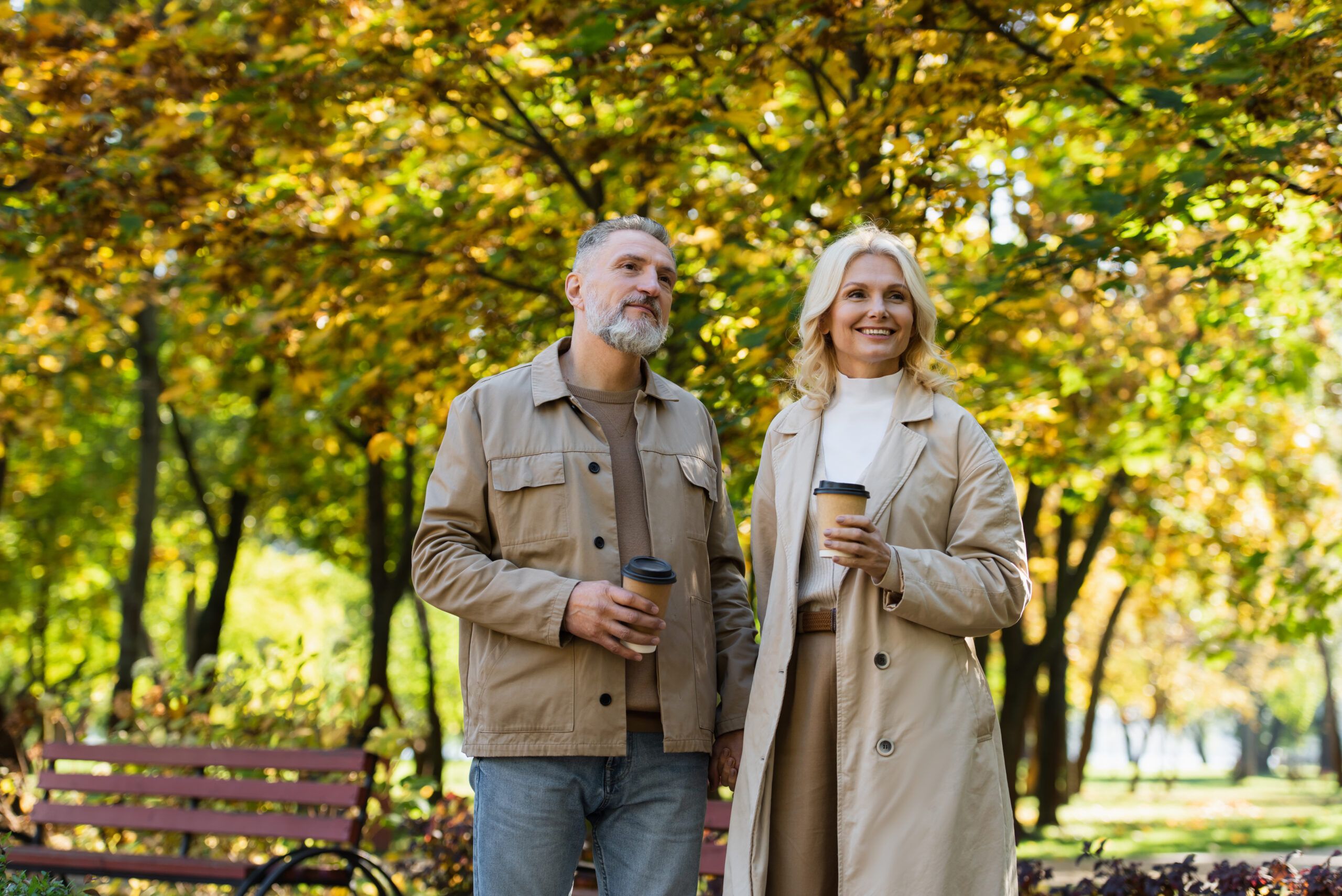 The Ensley 55+ Luxury Apartments with Smiling couple stands outdoors in a park with coffee cups, surrounded by autumn trees and sunlight.