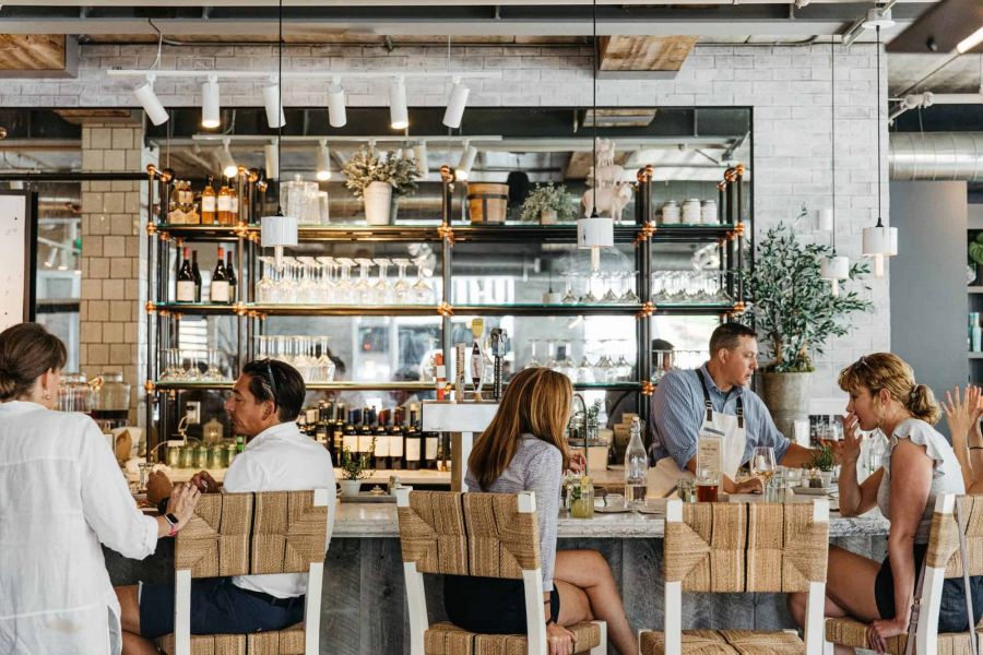 The Ensley 55+ Luxury Apartments with People sit at a modern restaurant bar, eating, drinking, and talking, with shelves of bottles and glasses behind them.