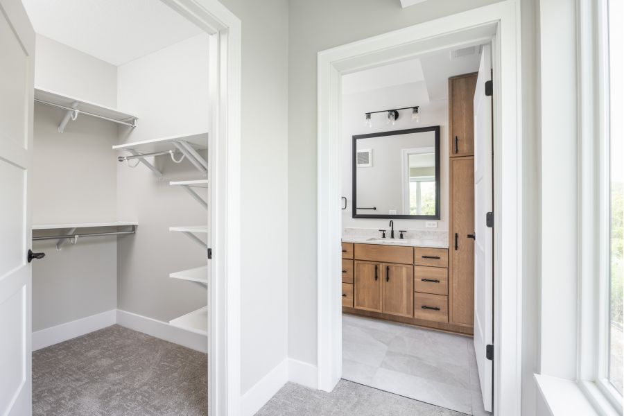 The Ensley 55+ Luxury Apartments with A walk-in closet with shelves is next to a modern bathroom with wood cabinets and a large mirror.