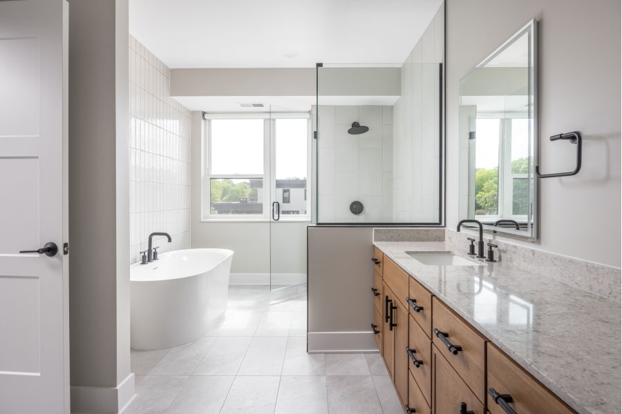 The Ensley 55+ Luxury Apartments with Modern bathroom with a freestanding tub, glass shower, large mirror, wood cabinets, and double sinks, all in neutral tones.