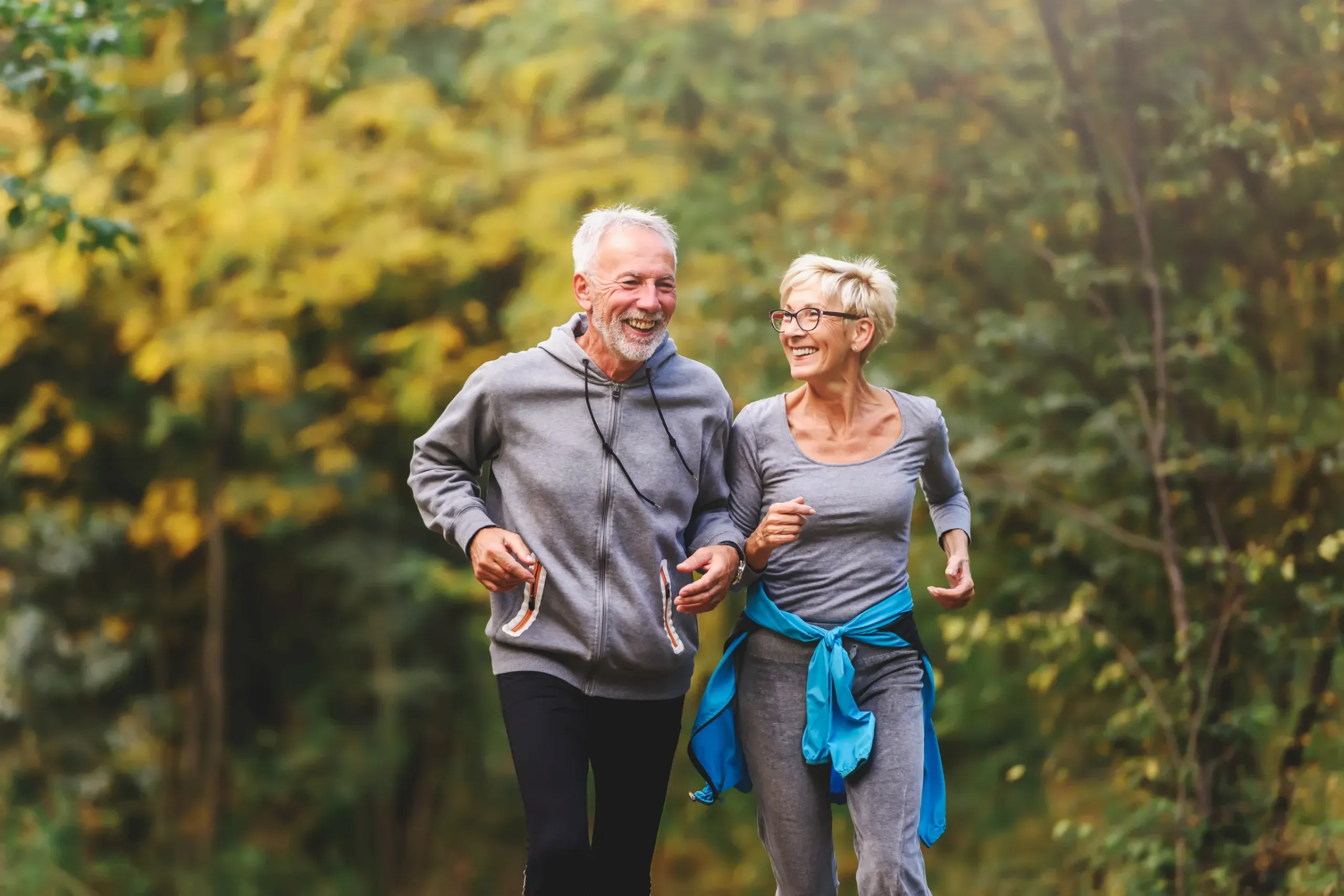 The Ensley 55+ Luxury Apartments with Older couple in activewear jogging and smiling together outdoors in a forest or park.