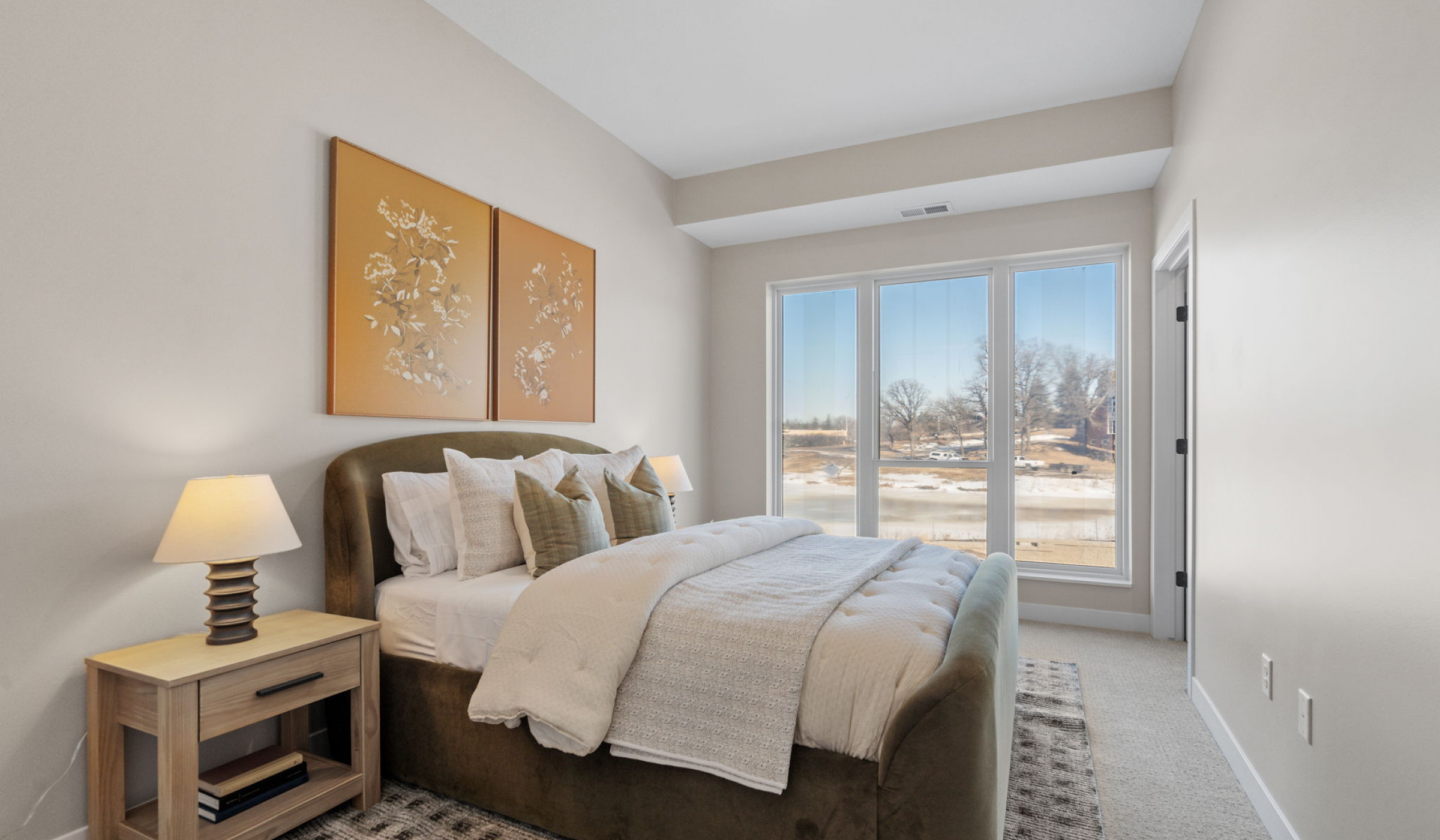 The Ensley 55+ Luxury Apartments with Bright bedroom with large windows, a cozy bed, nightstand, lamps, and wall art above the headboard.