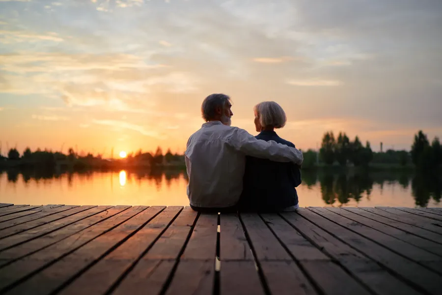The Ensley 55+ Luxury Apartments with An older couple sits on a wooden dock, watching the sunset over a calm lake.