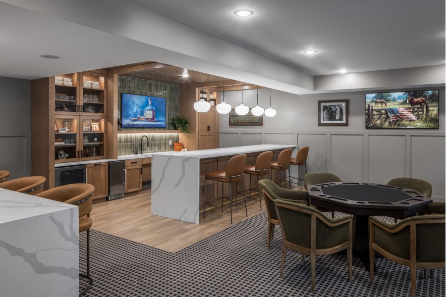 The Ensley 55+ Luxury Apartments with Modern kitchen and bar with marble countertops, brown chairs, poker table, and art on the walls under bright lighting.