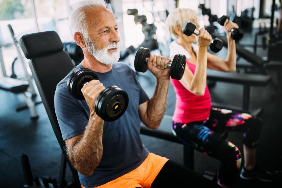 The Ensley 55+ Luxury Apartments with Two older adults lift dumbbells together while seated on benches in a gym, focused on their workout.