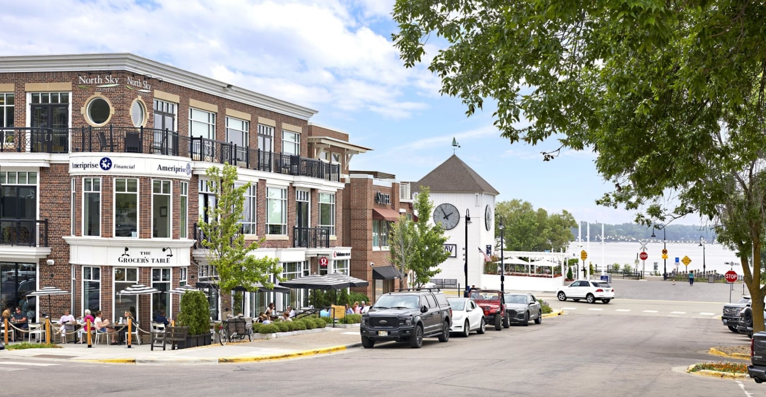 The Ensley 55+ Luxury Apartments with Street view of a lakeside town with shops, parked cars, outdoor dining, and a white clock tower.
