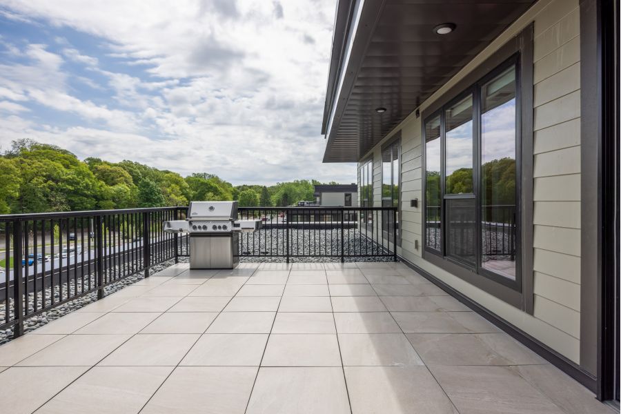 The Ensley 55+ Luxury Apartments with Spacious rooftop patio with tile flooring, black railing, and a stainless steel grill, overlooking green trees under a partly cloudy sky.