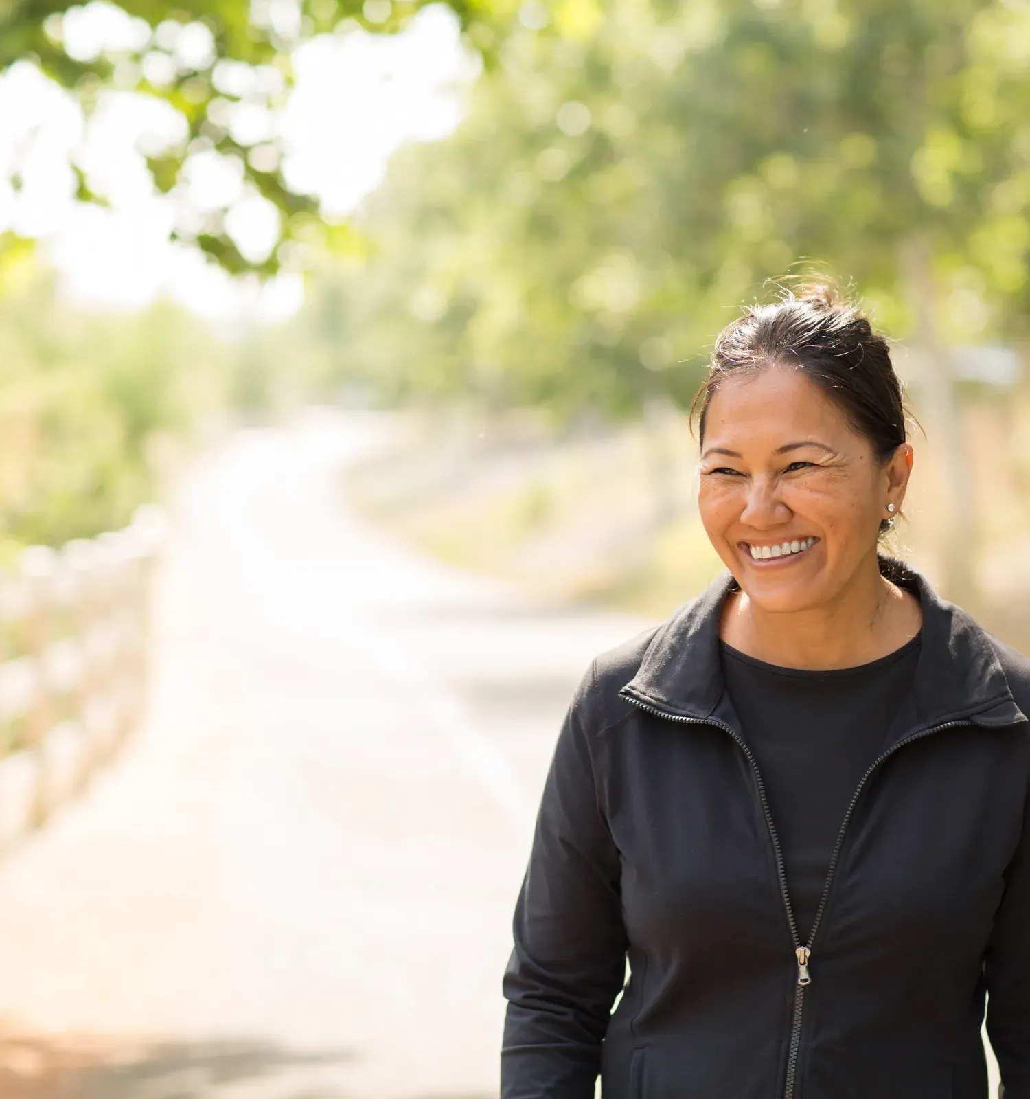 The Ensley 55+ Luxury Apartments with Woman smiling outdoors on a sunny day, standing on a path surrounded by trees and greenery.
