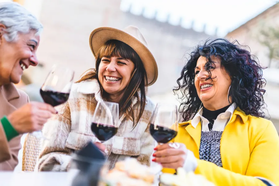 The Ensley 55+ Luxury Apartments with Three women laughing and clinking glasses of red wine while sitting together outdoors.