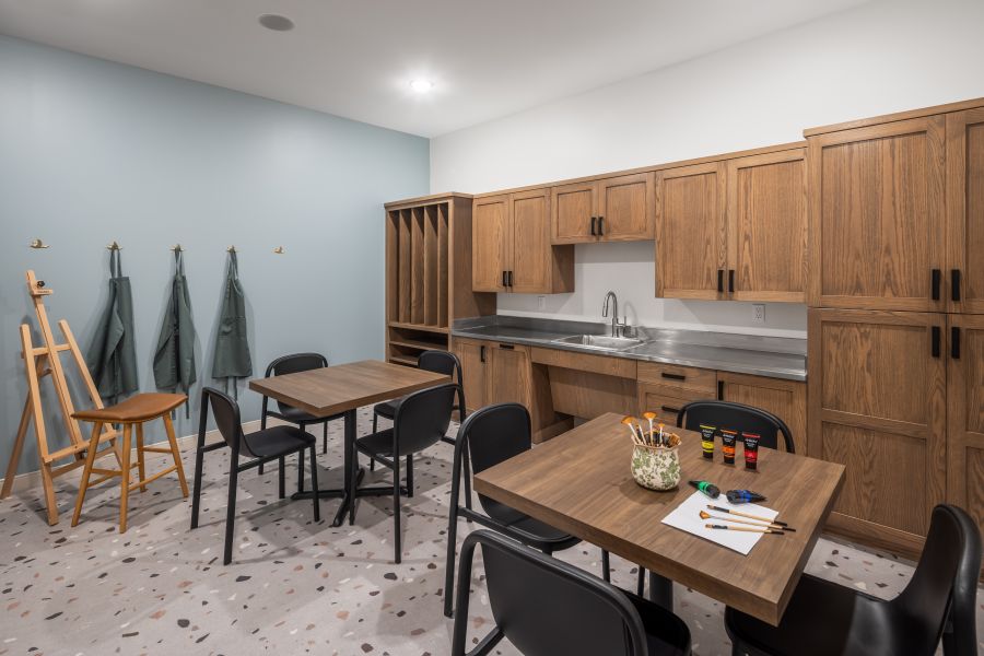 The Ensley 55+ Luxury Apartments with Art classroom with wooden cabinets, tables, chairs, aprons on hooks, an easel, art supplies, and a sink along the back wall.