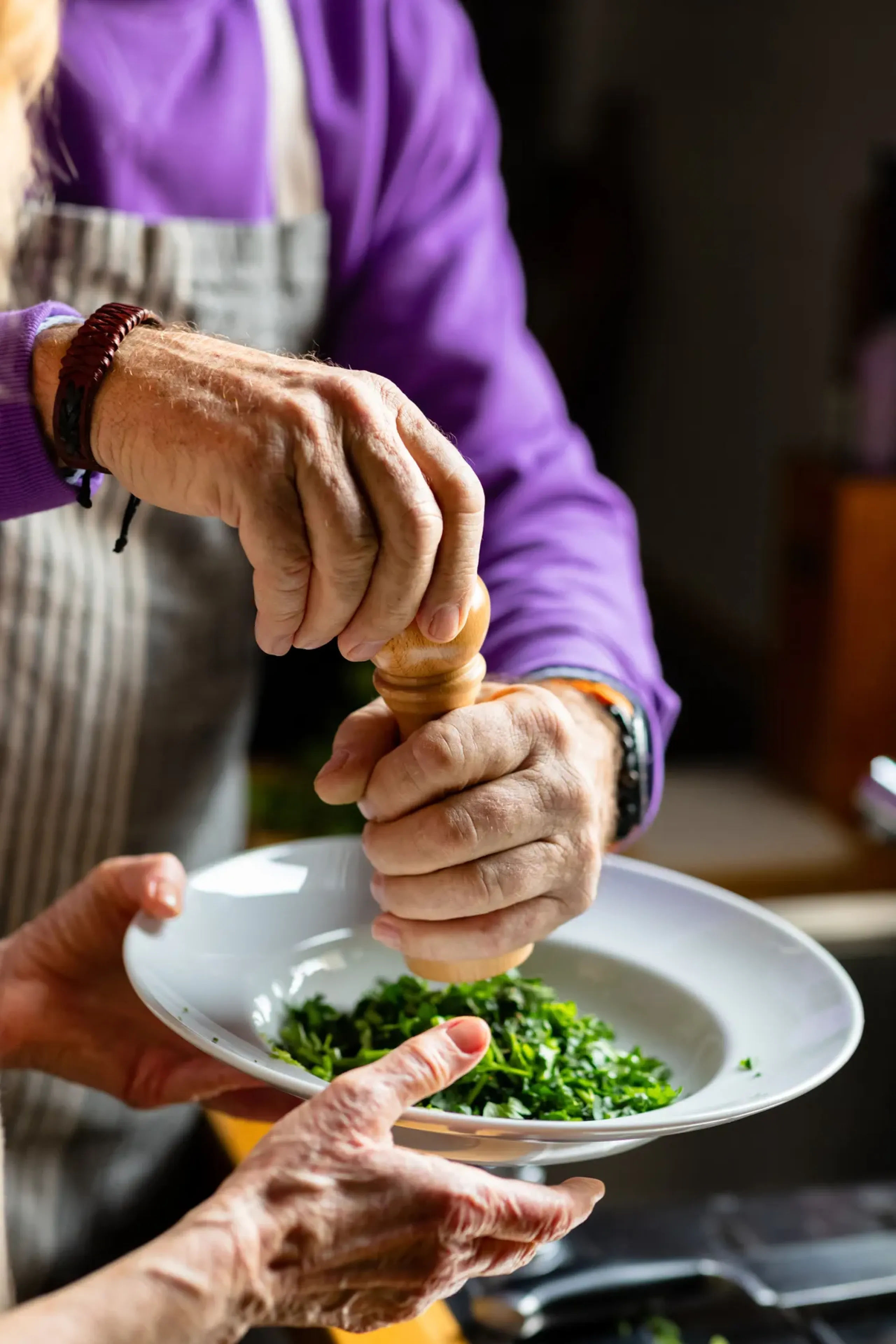 The Ensley 55+ Luxury Apartments with Person grinding pepper onto chopped herbs in a white bowl, held by another person in the kitchen.