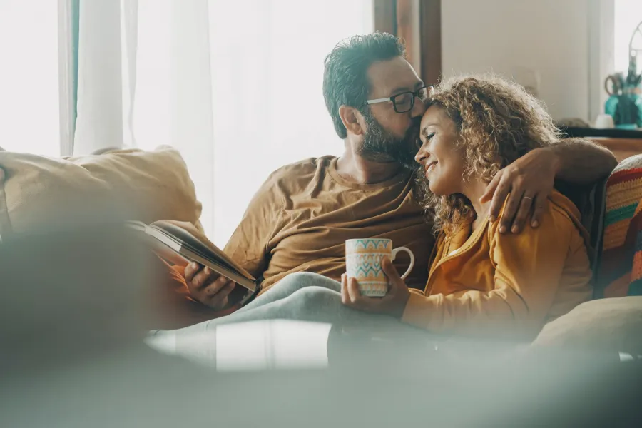 The Ensley 55+ Luxury Apartments with A man kisses a woman's forehead as they cuddle on a couch; she holds a mug and he holds a book.