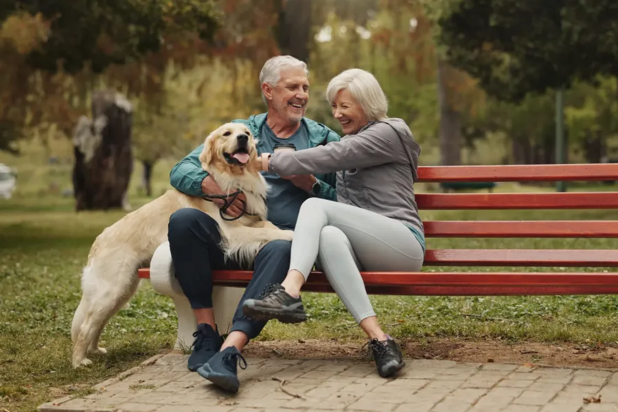 The Ensley 55+ Luxury Apartments with Smiling older couple sitting on a park bench with their golden retriever dog.